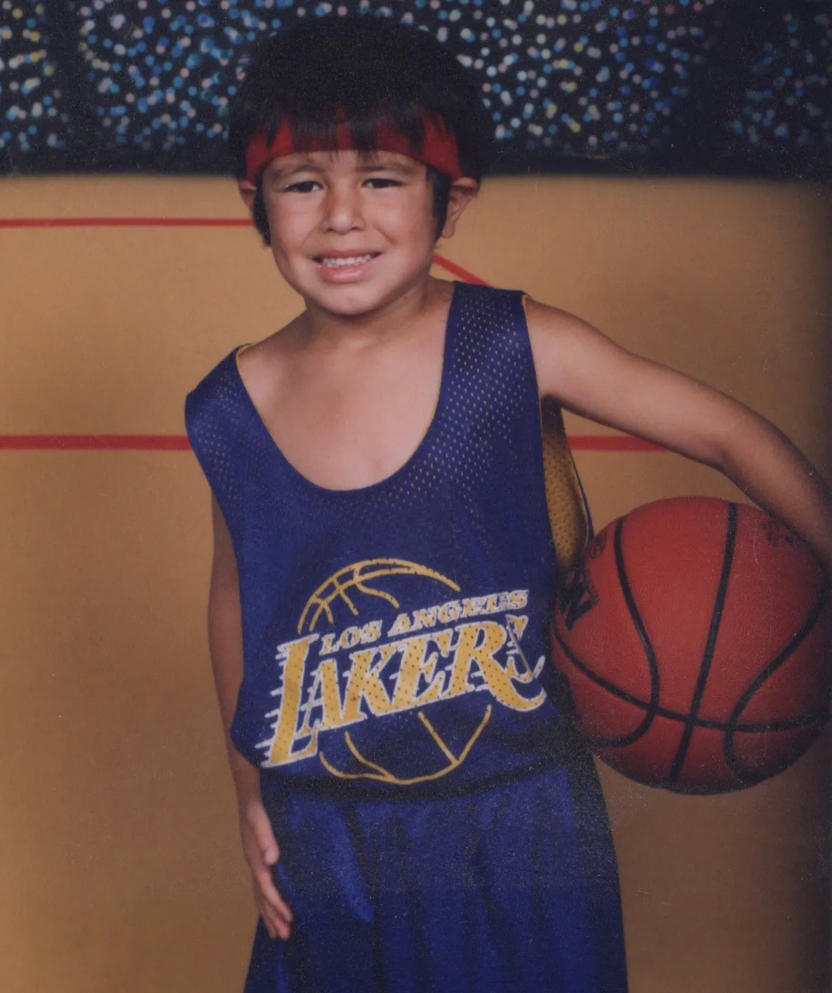 A young Mike De La Rosa in a Los Angeles Lakers jersey and headband, holding a basketball in a youth-league team portrait.
